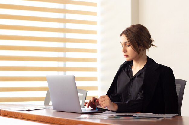 A front view young beautiful businesswoman in black shirt black jacket using her silver laptop writing reading working inside her office work job building