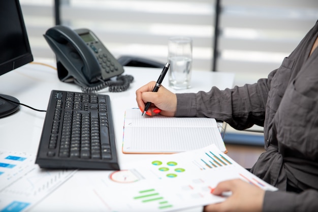 A front view young beautiful businesswoman working on her pc on the table along with phone and graphics writing down notes job activities technology