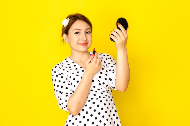 A front view young beautiful female in black and white polka dot dress doing make-up on yellow background clothing fashion mascara brush lipstick
