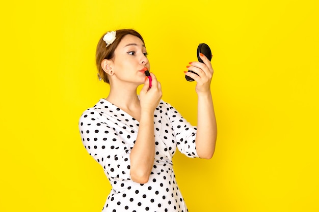 Una vista frontale giovane bella femmina in abito bianco e nero a pois facendo il trucco su sfondo giallo abbigliamento moda mascara rossetto pennello