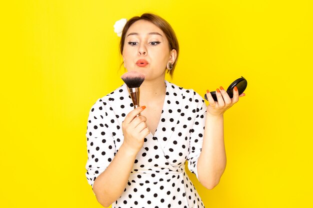 A front view young beautiful female in black and white polka dot dress doing make-up on yellow