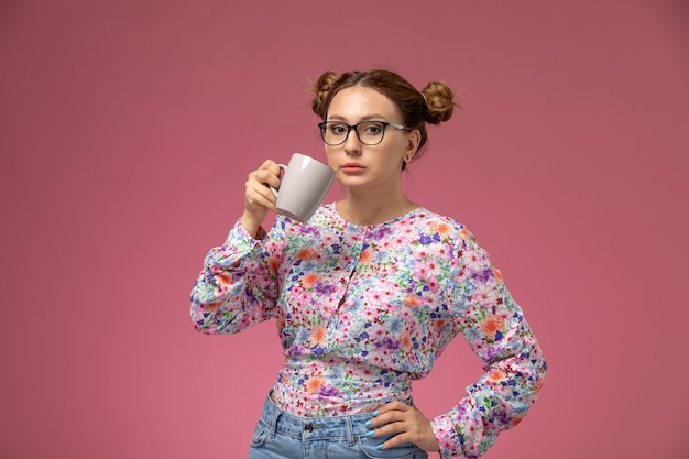 Front view young beautiful female in flower designed shirt and blue jeans drinking tea posing on the pink background