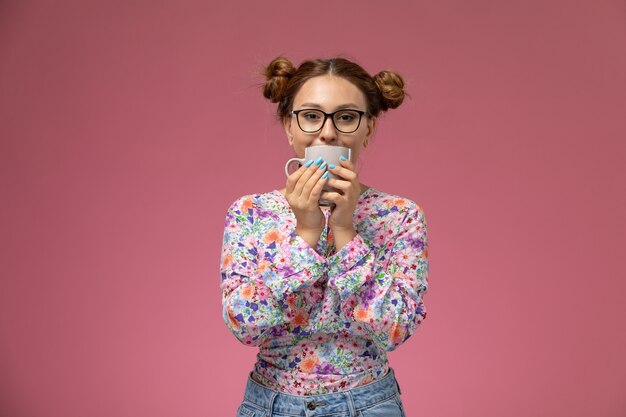 Front view young beautiful female in flower designed shirt and blue jeans drinking a tea smiling on pink background