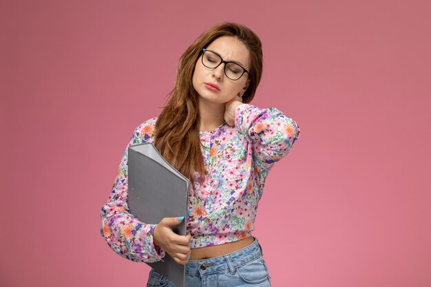 Front view young beautiful female in flower designed shirt and blue jeans having neck ache on the pink background