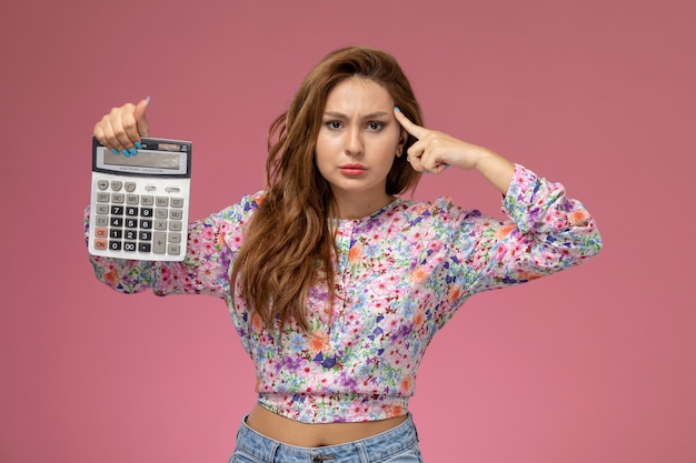 Front view young beautiful female in flower designed shirt and blue jeans holding calculator on pink background