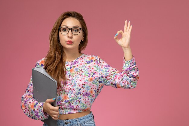 Front view young beautiful female in flower designed shirt and blue jeans holding grey colored document on pink background