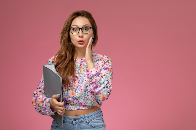 Front view young beautiful female in flower designed shirt and blue jeans holding grey document on pink background