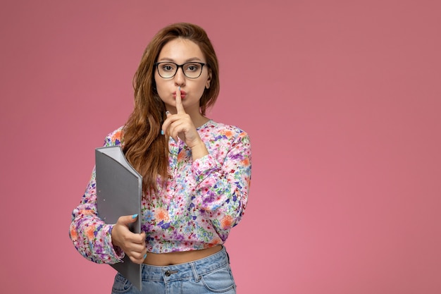 Front view young beautiful female in flower designed shirt and blue jeans holding grey file showing silence sign on the pink background