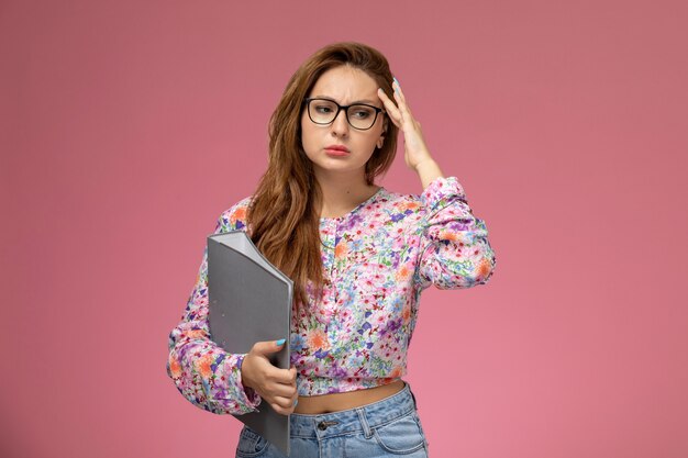 Front view young beautiful female in flower designed shirt and blue jeans holding grey file with depressed expression on the pink background