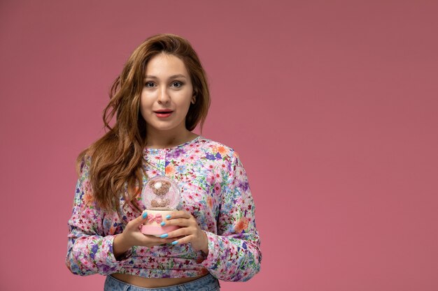 Front view young beautiful female in flower designed shirt and blue jeans holding little glass toy on the pink background