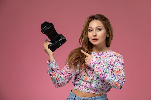 Free photo front view young beautiful female in flower designed shirt and blue jeans holding a photo camera on pink background