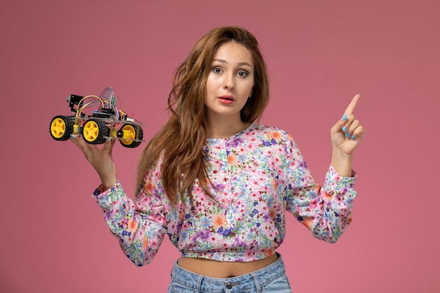 Front view young beautiful female in flower designed shirt and blue jeans holding toy car posing on pink background