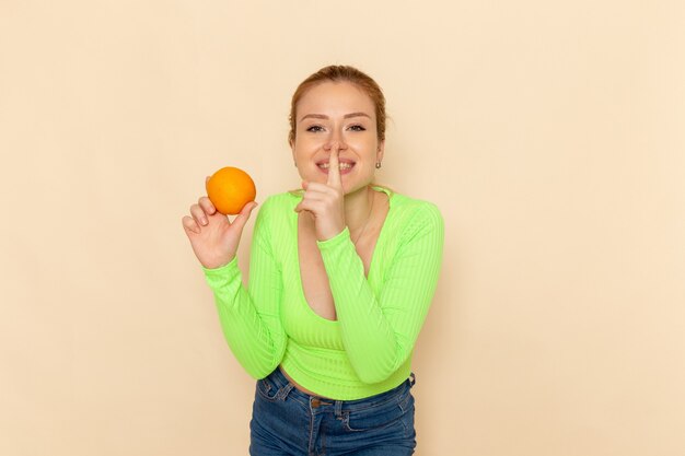 クリーム色の壁のフルーツモデルの女性のまろやかな笑顔で新鮮なオレンジ色のシャツを保持している緑のシャツの若い美しい女性の正面図