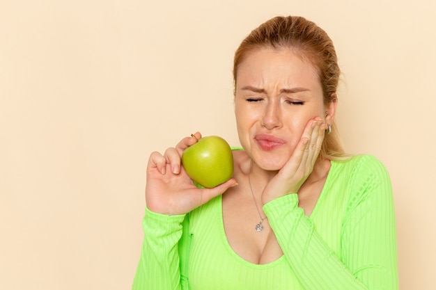 Вид спереди молодая красивая женщина в зеленой рубашке, держащая зеленое яблоко и имеющая зубную боль на кремовой стене, модель фруктов, женщина спелая
