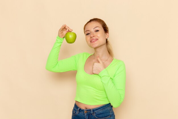 Front view young beautiful female in green shirt holding green fresh apple on cream desk fruits model woman mellow