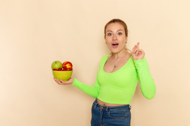 Front view young beautiful female in green shirt holding plate full of fruits on light-cream wall fruit model woman pose