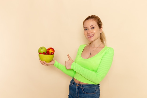 ライトクリーム色の壁に果物でいっぱいのプレートを保持している緑のシャツの正面図若い美しい女性フルーツモデルの女性のポーズ