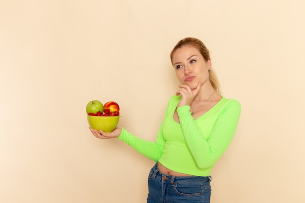 Front view young beautiful female in green shirt holding plate full of fruits thinking on the light cream wall fruit model woman pose