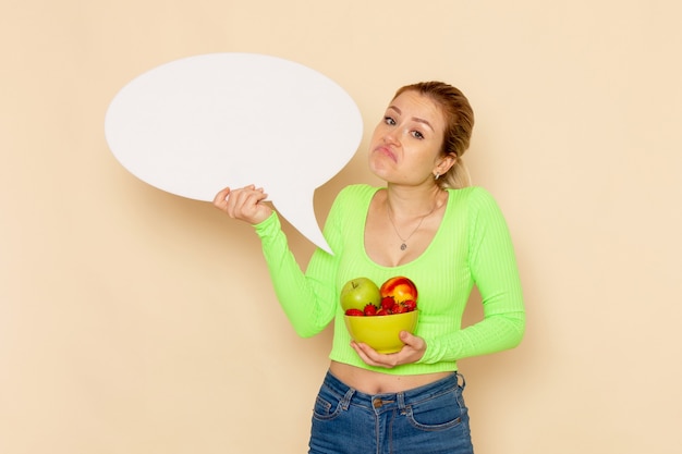 Front view young beautiful female in green shirt holding plate full of fruits and white sign on the cream wall fruit model woman food vitamine color