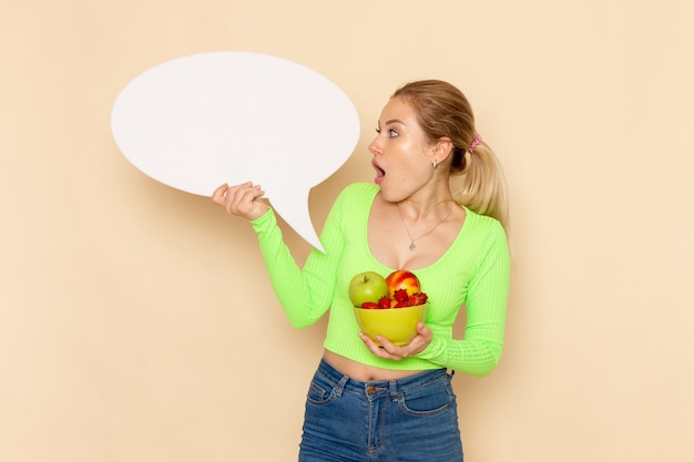 Front view young beautiful female in green shirt holding plate full of fruits and white sign on cream wall fruit model woman food