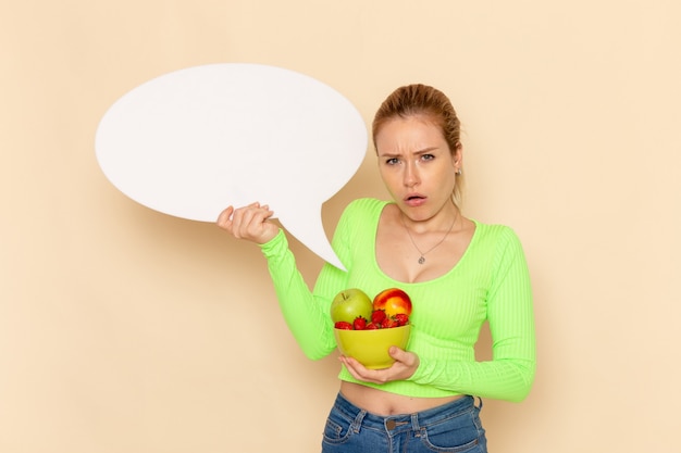 Front view young beautiful female in green shirt holding plate full of fruits and white sign on cream wall fruits model woman food vitamine color