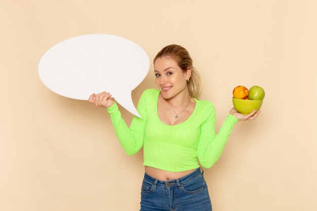 Вид спереди молодая красивая женщина в зеленой рубашке, держащая тарелку, полную фруктов с белым знаком на кремовой стене, модель фруктов, женщина, витамин