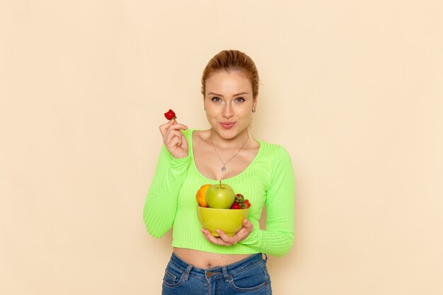 Вид спереди молодая красивая женщина в зеленой рубашке, держащая тарелку с фруктами, ест клубнику на светлой кремовой стене, фруктовая модель позы женщины