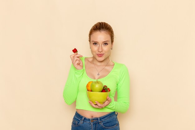 Front view young beautiful female in green shirt holding plate with fruits and eating strawberry on the light-cream wall fruit model woman pose