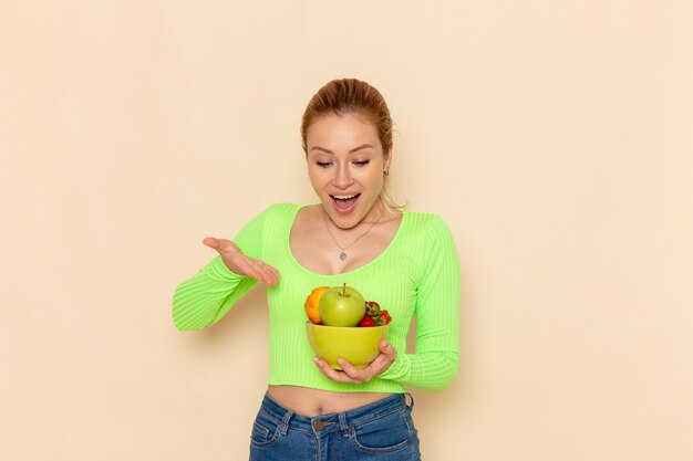 Front view young beautiful female in green shirt holding plate with fruits smiling on the light cream desk fruit model woman pose lady