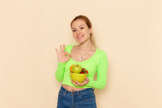Вид спереди молодая красивая женщина в зеленой рубашке, держащая тарелку с фруктами, улыбающаяся на светлом кремовом полу, модель фруктов, женщина, поза леди