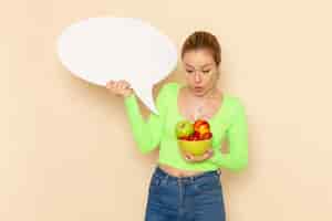 Бесплатное фото Вид спереди молодая красивая женщина в зеленой рубашке, держащая тарелку, полную фруктов и белый знак на кремовой стене, модель фруктов, женщина, пища, витамин