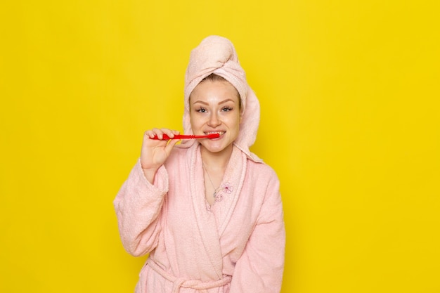 A front view young beautiful female in pink bathrobe cleaning her teeth