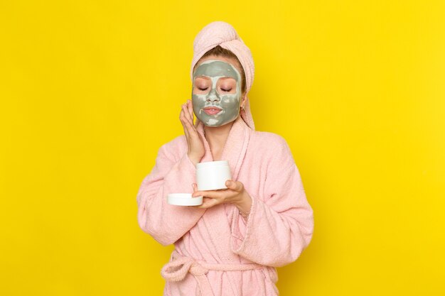 A front view young beautiful female in pink bathrobe holding cream can and using it