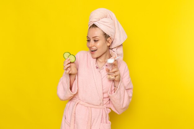 A front view young beautiful female in pink bathrobe holding cucumber rings and flask