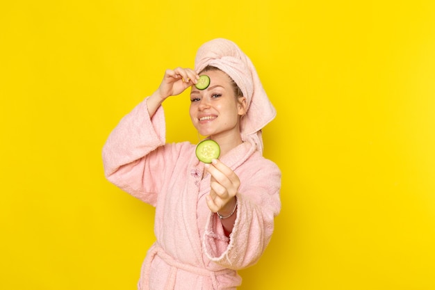 A front view young beautiful female in pink bathrobe holding cucumber slices and smiling