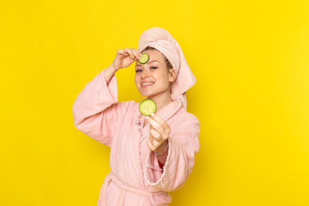 A front view young beautiful female in pink bathrobe holding cucumber slices and smiling