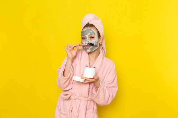A front view young beautiful female in pink bathrobe holding face cream can and brush