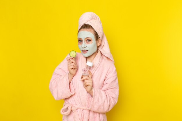 A front view young beautiful female in pink bathrobe holding flask and cucumber