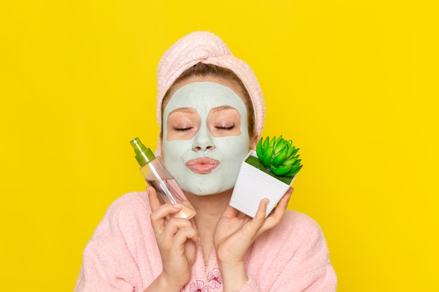 A front view young beautiful female in pink bathrobe holding make-up cleaner spray and little plant