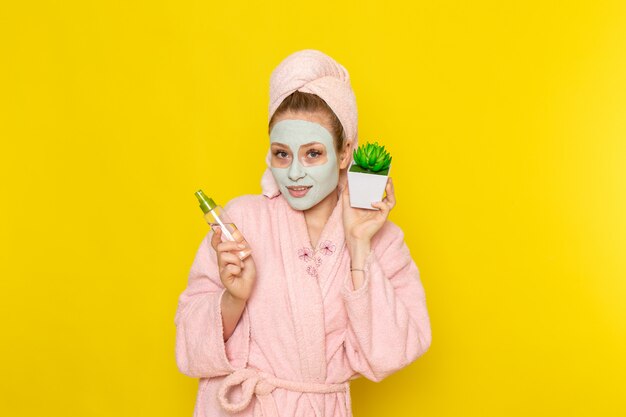 A front view young beautiful female in pink bathrobe holding make-up cleaner spray and little plant