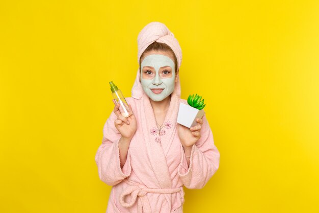 A front view young beautiful female in pink bathrobe holding make-up cleaner spray and little plant