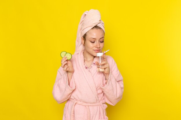 A front view young beautiful female in pink bathrobe holding spray and cucumber rounds