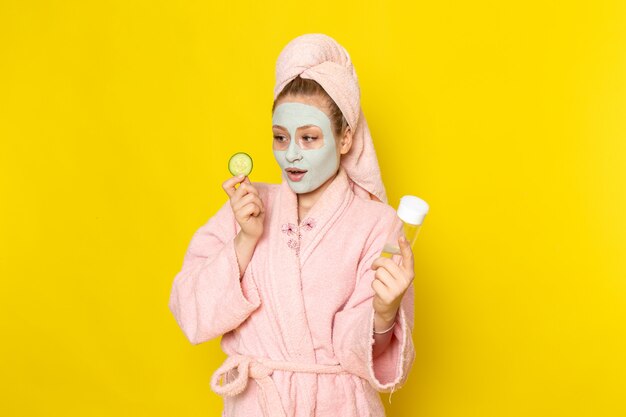 A front view young beautiful female in pink bathrobe holding spray and cucumber