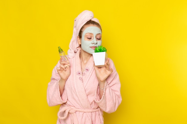 A front view young beautiful female in pink bathrobe holding spray flask and green plant on the yellow backgroun