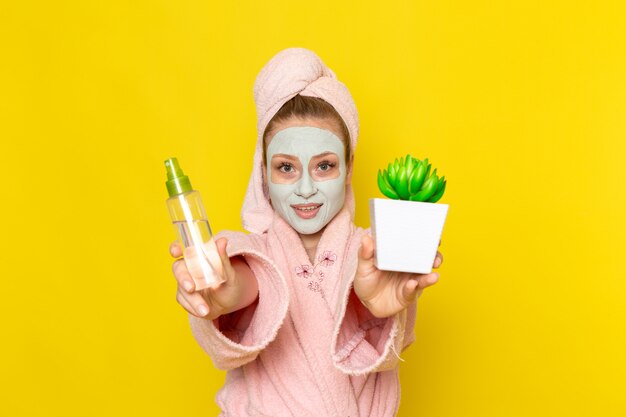 A front view young beautiful female in pink bathrobe holding spray and little green plant