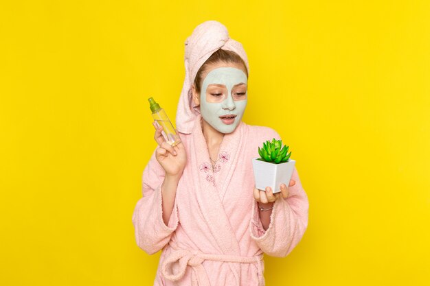 A front view young beautiful female in pink bathrobe holding spray and little plant