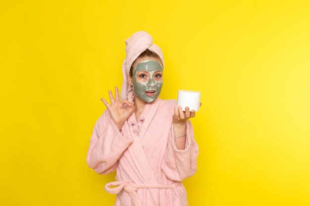 A front view young beautiful female in pink bathrobe with face-mask holding cream