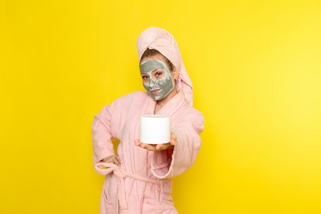 A front view young beautiful female in pink bathrobe with face-mask holding cream