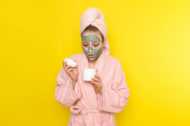 Free photo a front view young beautiful female in pink bathrobe with face-mask holding white cream