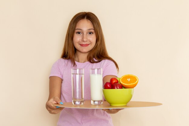 Вид спереди молодая красивая женщина в розовой футболке и синих джинсах, держащая поднос с фруктами, молоком и водой на сером